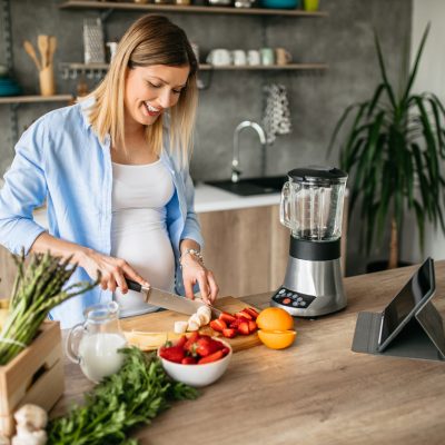 Young pregnant woman preparing fruit for healthy smoothie in her cute kitchen