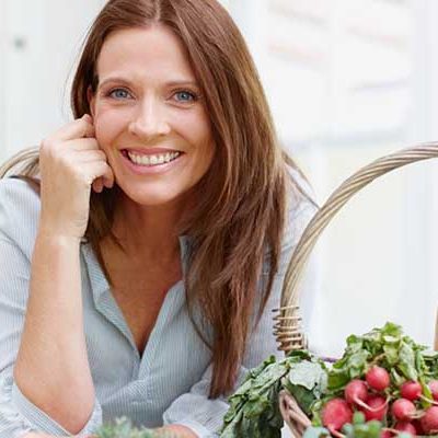 A beautiful woman leans on her kitchen counter behing a basket of fresh  vegetables